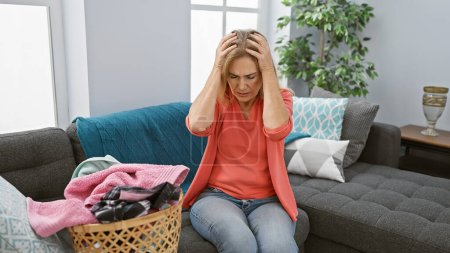 Photo for A stressed middle-aged woman sits on a couch with a laundry basket, feeling overwhelmed in her living room. - Royalty Free Image