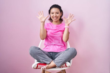Foto de Mujer joven hispana sentada en silla sobre fondo rosa mostrando y señalando hacia arriba con los dedos número diez mientras sonríe confiada y feliz. - Imagen libre de derechos