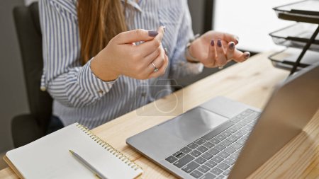 Photo for A young brunette woman with long hair focused on work in a modern office, holding a pen near a laptop. - Royalty Free Image