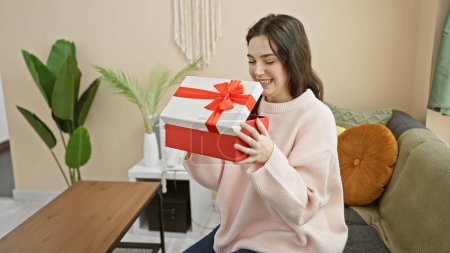 Téléchargez les photos : Une jeune femme souriante tenant une boîte-cadeau rouge à la maison, exsudant bonheur et chaleur dans un salon confortable. - en image libre de droit