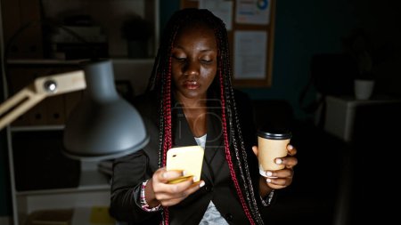 Téléchargez les photos : Femme afro-américaine ouvrière utilisant un smartphone buvant du café au bureau - en image libre de droit