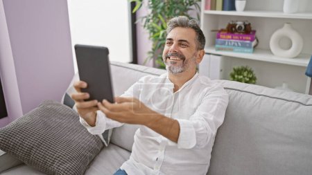 Foto de Alegre joven hispano con el pelo gris sonriendo y teniendo una divertida videollamada en touchpad, relajándose en su sofá de la sala de estar - Imagen libre de derechos