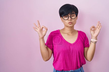 Téléchargez les photos : Jeune femme asiatique aux cheveux courts debout sur fond rose détendue et souriante avec les yeux fermés faisant geste de méditation avec les doigts. concept de yoga. - en image libre de droit