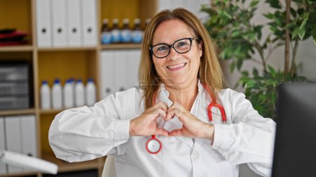 Téléchargez les photos : Moyen Age hispanique femme médecin souriant faire geste cardiaque avec les mains à la clinique - en image libre de droit
