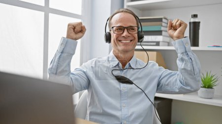 Photo for Cheerful middle age business man celebrates with a winning gesture at his office, enjoying catchy music on his laptop through headphones - Royalty Free Image