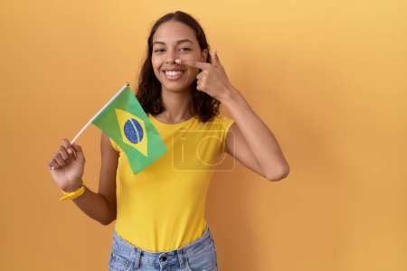 Téléchargez les photos : Jeune femme hispanique tenant le drapeau brésilien pointant du doigt la main vers le visage et le nez, souriante et joyeuse. concept de beauté - en image libre de droit