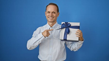 Photo for Cheerful middle age man beaming with joy and confidence, pointing to a surprising birthday gift against an isolated white background, his positive expression contagiously spreading happiness - Royalty Free Image