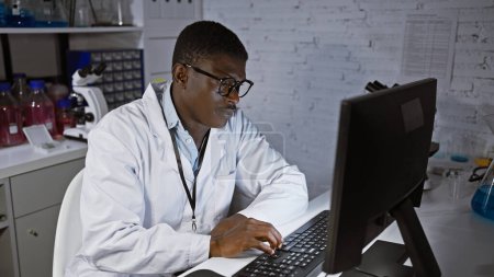 Téléchargez les photos : Homme africain en blouse de laboratoire travaillant sur ordinateur dans un cadre de laboratoire avec des équipements scientifiques - en image libre de droit