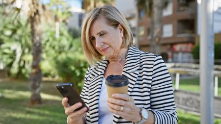 Foto de Mujer madura sosteniendo café y teléfono inteligente al aire libre en un parque - Imagen libre de derechos
