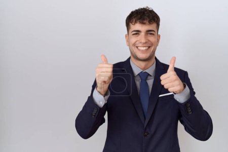 Téléchargez les photos : Jeune homme d'affaires hispanique portant costume et cravate signe de succès faire geste positif avec la main, pouces levés souriant et heureux. expression joyeuse et geste gagnant. - en image libre de droit
