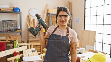 Photo for A smiling young woman in a workshop holding a drill portrays skilled carpentry indoors. - Royalty Free Image