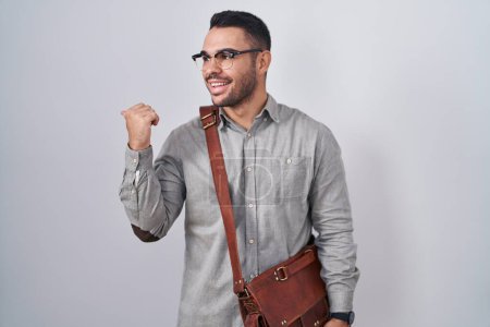 Téléchargez les photos : Jeune homme hispanique portant une valise souriant avec le visage heureux regardant et pointant vers le côté avec le pouce vers le haut. - en image libre de droit