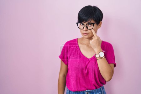 Foto de Joven mujer asiática con pelo corto de pie sobre fondo rosa apuntando al ojo observándote gesto, expresión sospechosa - Imagen libre de derechos