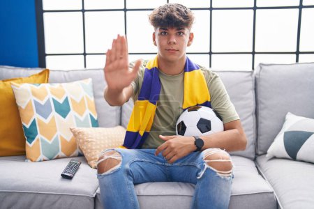 Téléchargez les photos : Adolescent hispanique assis sur le canapé regarder match de football avec la main ouverte faire stop signe avec expression sérieuse et confiante, geste de défense - en image libre de droit