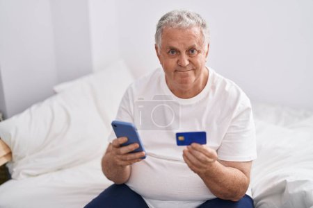 Foto de Hombre de pelo gris de mediana edad usando teléfono inteligente y tarjeta de crédito sentado en la cama en el dormitorio - Imagen libre de derechos