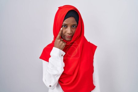 Téléchargez les photos : Jeune femme arabe portant l'écharpe traditionnelle islamique hijab pointant vers l'œil vous regardant geste, expression suspecte - en image libre de droit
