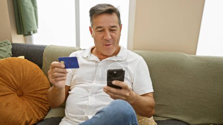 Middle-aged man holding credit card and smartphone on a couch indoors, possibly shopping online or managing finances at home