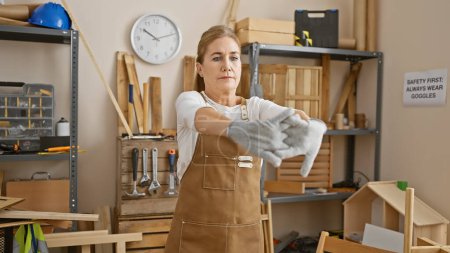 Photo for Mature woman in apron stretching gloves in a woodworking studio indicates safety and skill. - Royalty Free Image