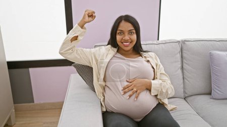 Foto de Mujer embarazada joven tocando el vientre haciendo un gesto fuerte con el brazo sonriendo en casa - Imagen libre de derechos
