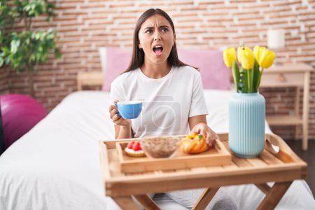 Foto de Joven mujer hispana comiendo pasteles para desayunar sentada en la cama enojada y loca gritando frustrada y furiosa, gritando con ira mirando hacia arriba. - Imagen libre de derechos