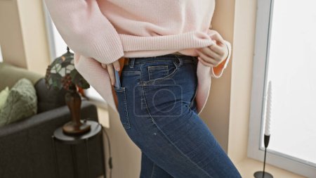 Téléchargez les photos : Une jeune femme caucasienne stocke avec désinvolture un smartphone dans sa poche de jeans dans un salon confortable. - en image libre de droit