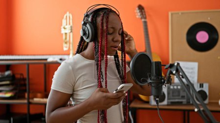 Foto de Sincera melodía, mujer afroamericana músico cantando conmovedoramente, mirando a la pantalla del teléfono inteligente en el estudio de música - Imagen libre de derechos