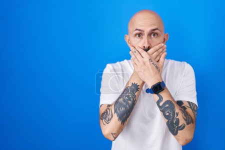 Téléchargez les photos : Homme hispanique avec des tatouages debout sur fond bleu choqué couvrant la bouche avec les mains pour erreur. concept secret. - en image libre de droit