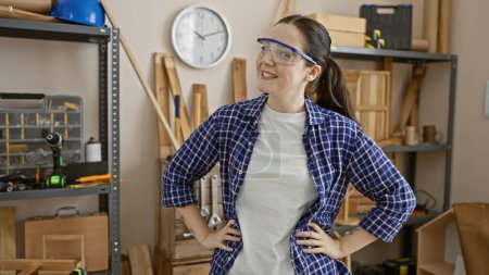 Photo for Smiling caucasian woman wearing safety glasses stands confidently in a well-organized carpentry workshop. - Royalty Free Image