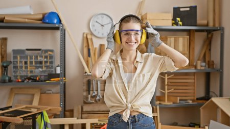 Photo for A smiling young woman wearing safety goggles and ear protection in a well-equipped carpentry workshop. - Royalty Free Image