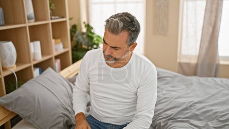Photo for Handsome, young hispanic man with grey hair, showing a serious expression while sitting on a comfortable bed and intensely thinking, engulfed in worry, in the relaxed indoors of his apartment bedroom. - Royalty Free Image