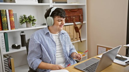 Téléchargez les photos : Jeune homme aime la musique avec écouteurs tout en travaillant sur ordinateur portable dans le bureau moderne - en image libre de droit