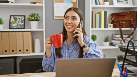 Foto de Una joven hispana sonriente se involucra en una conversación telefónica mientras sostiene una taza roja en un entorno de oficina moderno. - Imagen libre de derechos