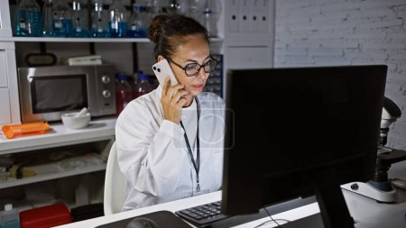 Photo for A mature hispanic woman in a lab coat talks on the phone while working on a computer in a laboratory setting. - Royalty Free Image