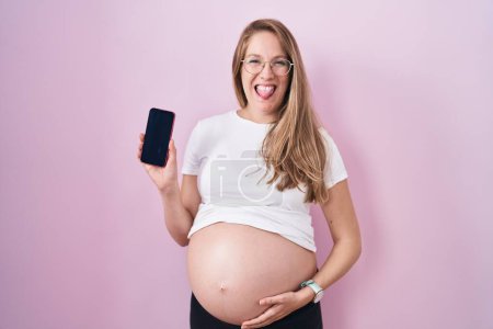 Téléchargez les photos : Jeune femme enceinte montrant ventre tenant smartphone langue collante heureux avec une expression drôle. - en image libre de droit