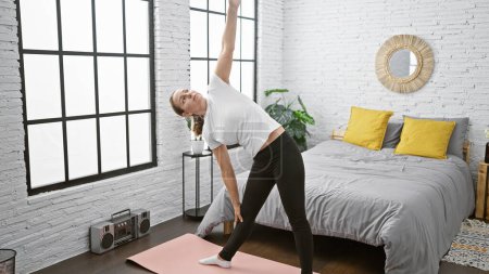 Téléchargez les photos : Serieux jeune femme blonde trouver l'équilibre, renforcer sa concentration avec l'exercice de yoga du matin sur son lit, échauffement dans un espace confortable chambre à coucher intérieure, profiter du calme et de détente. - en image libre de droit