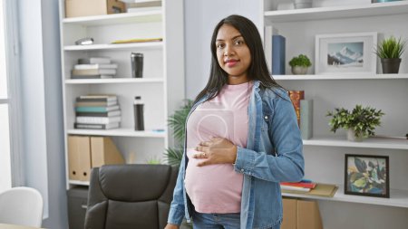 Foto de Sonriente joven embarazada toca con confianza el vientre mientras trabaja en la oficina, encarnando la maternidad y el éxito empresarial. - Imagen libre de derechos