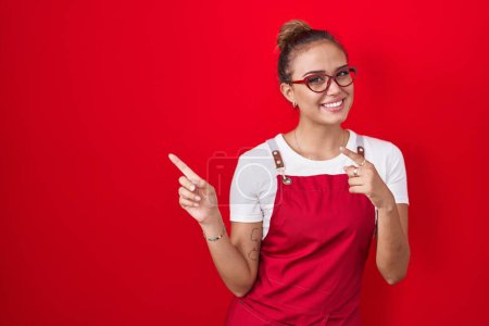 Photo for Young hispanic woman wearing waitress apron over red background smiling and looking at the camera pointing with two hands and fingers to the side. - Royalty Free Image