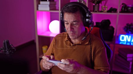 Photo for A middle-aged man wearing headphones examines a smartphone in a modern, dimly-lit gaming room at night. - Royalty Free Image