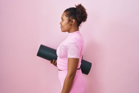 Photo for Young hispanic woman with curly hair holding yoga mat over pink background looking to side, relax profile pose with natural face and confident smile. - Royalty Free Image