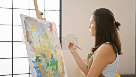 Photo for Thoughtful young beautiful hispanic woman artist, concentrating and doubting with the brush in hand, looking at her drawing on the easel in the art studio classroom. - Royalty Free Image