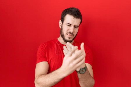 Foto de Hombre hispano joven con camiseta roja casual que sufre dolor en las manos y los dedos, inflamación de la artritis - Imagen libre de derechos