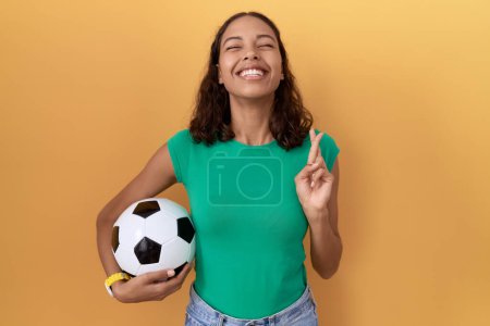Foto de Mujer hispana joven sosteniendo la pelota señalando con el dedo cruzada sonriendo con esperanza y los ojos cerrados. suerte y concepto supersticioso. - Imagen libre de derechos