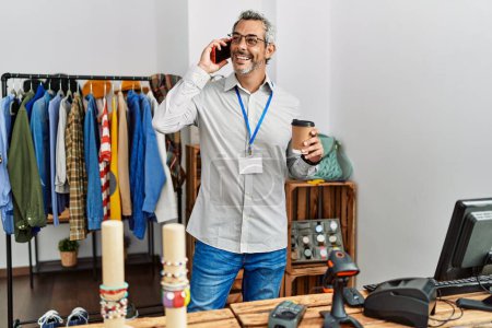 Foto de Asistente de tienda hombre de pelo gris de mediana edad hablando en el teléfono inteligente beber café en la tienda de ropa - Imagen libre de derechos