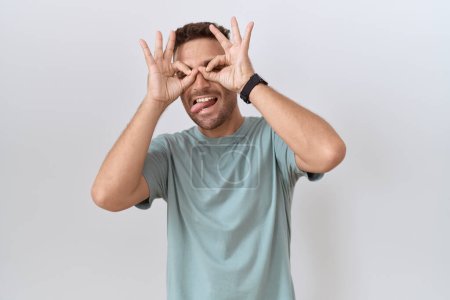Foto de Hombre hispano con barba de pie sobre fondo blanco haciendo buen gesto como binoculares sacando la lengua, ojos mirando a través de los dedos. expresión loca. - Imagen libre de derechos