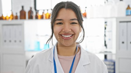 Photo for A smiling young hispanic woman in a white lab coat standing in a pharmacy. - Royalty Free Image