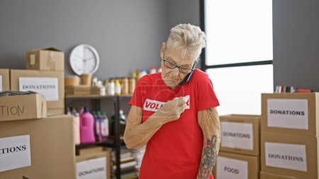 Téléchargez les photos : Une femme âgée, travailleuse et aux cheveux gris, bénévole engagée dans une conversation téléphonique sérieuse au centre communautaire de bienfaisance, entourée de boîtes de dons - en image libre de droit