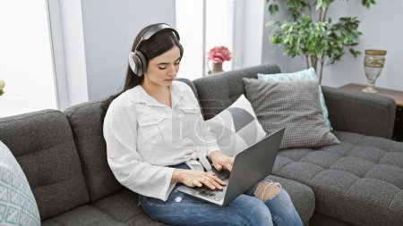 Una mujer hispana enfocada usando auriculares usa una computadora portátil mientras está sentada en un sofá en su sala de estar.