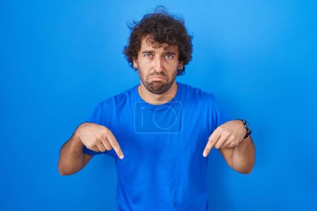 Photo for Hispanic young man standing over blue background pointing down looking sad and upset, indicating direction with fingers, unhappy and depressed. - Royalty Free Image