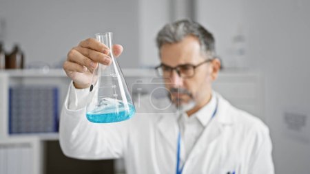 Photo for Attractive young hispanic man, grey-haired chemist, concentrating on measuring liquid in test tube at indoor science lab. - Royalty Free Image