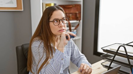 Photo for Pensive woman in glasses at her desk in a modern office, embodying professionalism and concentration - Royalty Free Image
